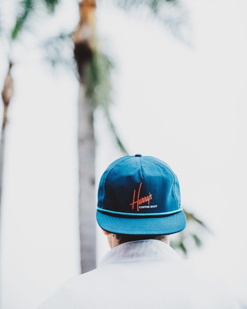 Young man walking away wearing a blue Harry's Coffee Shop hat backwards.