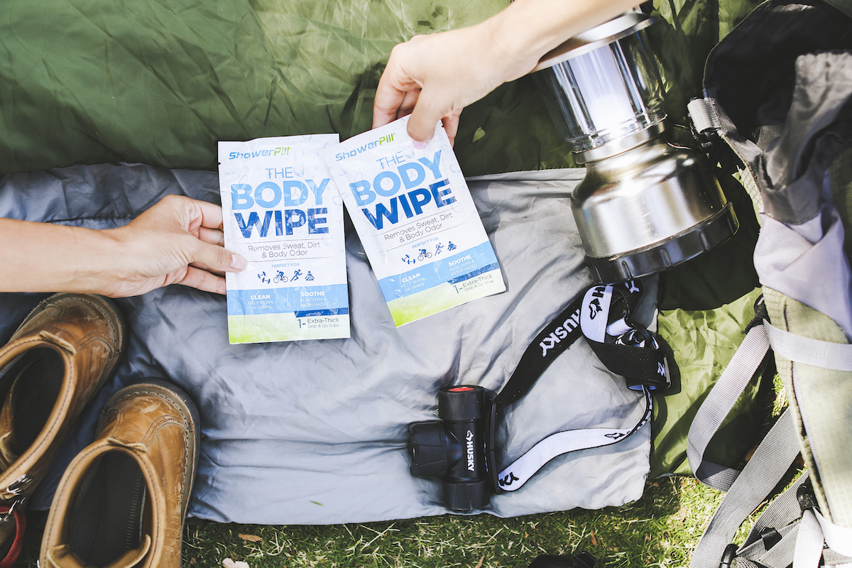 Two hands reaching for two ShowerPill body wipes set amongst camping equipment