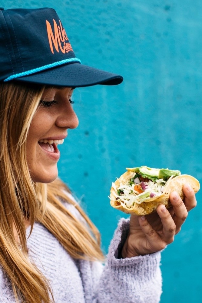 A young woman wearing a Mike's Taco Club Hat eating a taco.